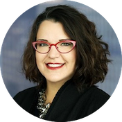 A woman with shoulder-length dark hair, wearing red glasses and a black blazer, smiling at the camera against a blue background.