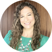 Portrait of a smiling woman with curly hair wearing a teal top with a decorative necklace, standing against a wooden background.