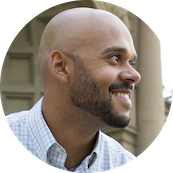 A smiling man with a shaved head and beard wearing a light blue checkered shirt, standing outdoors in front of a stone building.