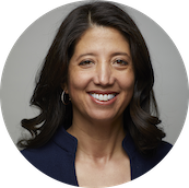 A woman with dark brown hair smiling, wearing a navy blazer and earrings, against a neutral background.