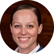 Close-up of a smiling woman with short brown hair and blue eyes, wearing a dark blazer, against a dark wooden background.