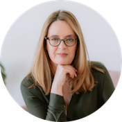A woman with glasses and long blonde hair, resting her chin on her hand, looking at the camera