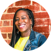 Smiling woman with braids, wearing a denim jacket and yellow top, standing in front of a brick wall.