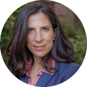 A woman with long dark hair, wearing a blue blazer and pink shirt, outdoors with greenery in the background.