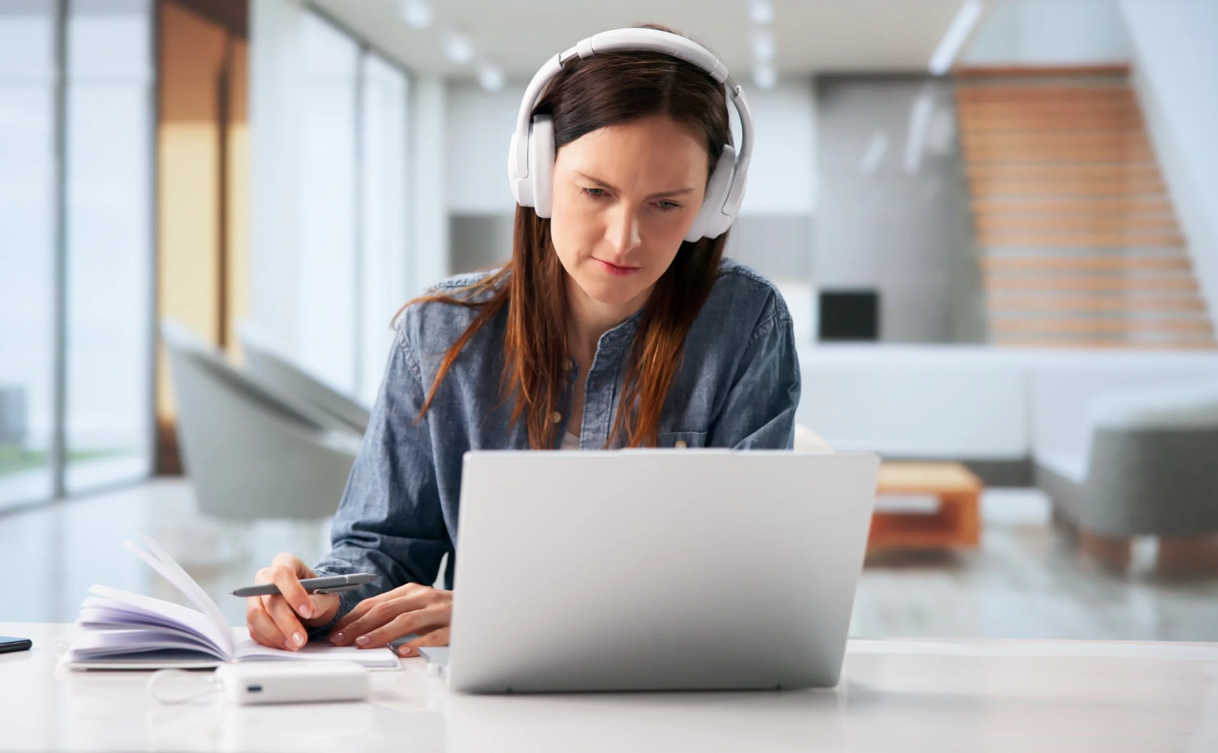 A woman sitting at a table, wearing headphones, working on a laptop with an open notebook and pen in hand, in a modern, well-lit indoor setting.