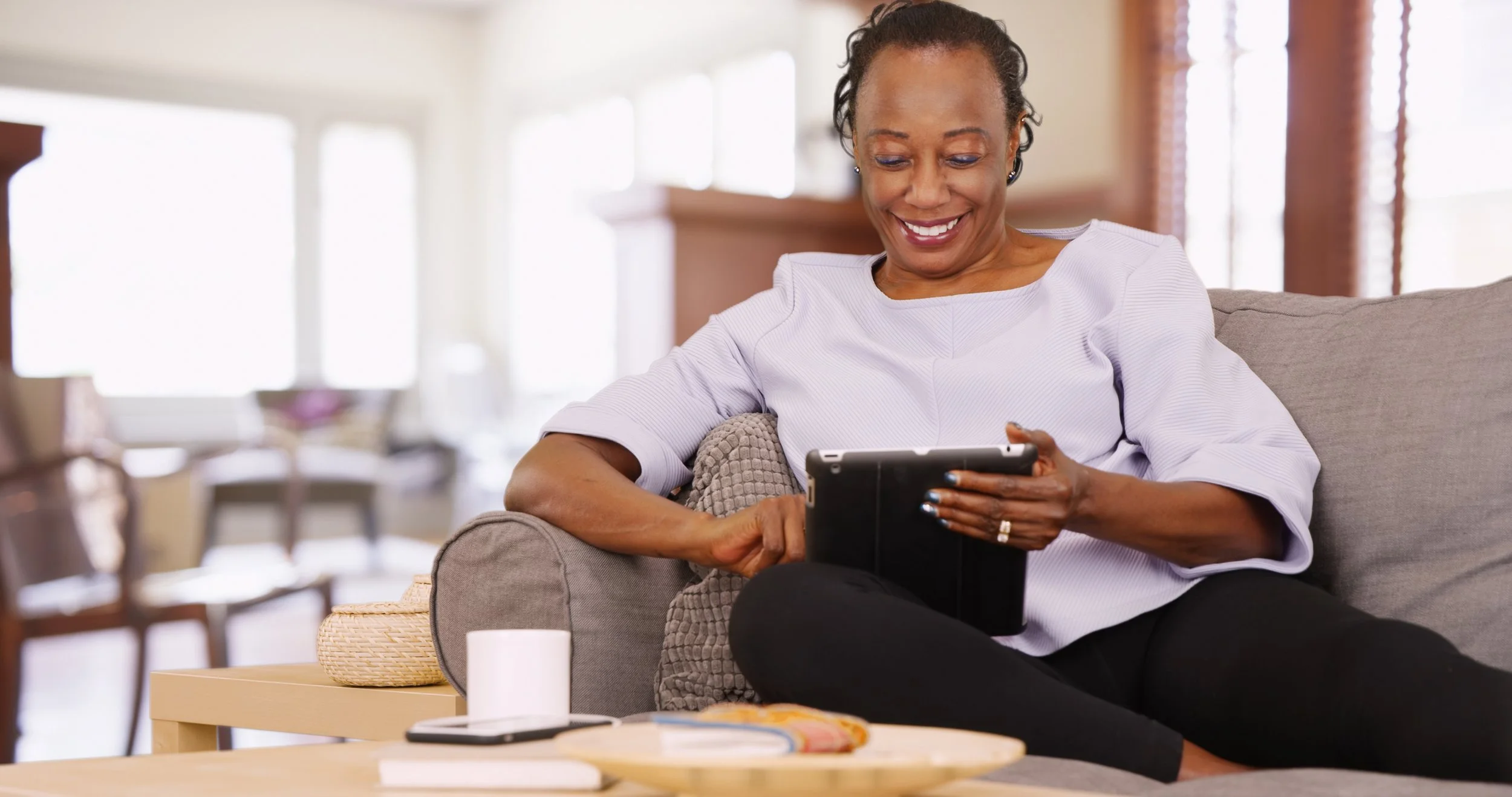 A smiling woman sitting on a gray couch, looking at her smartphone in a bright living room with large windows.