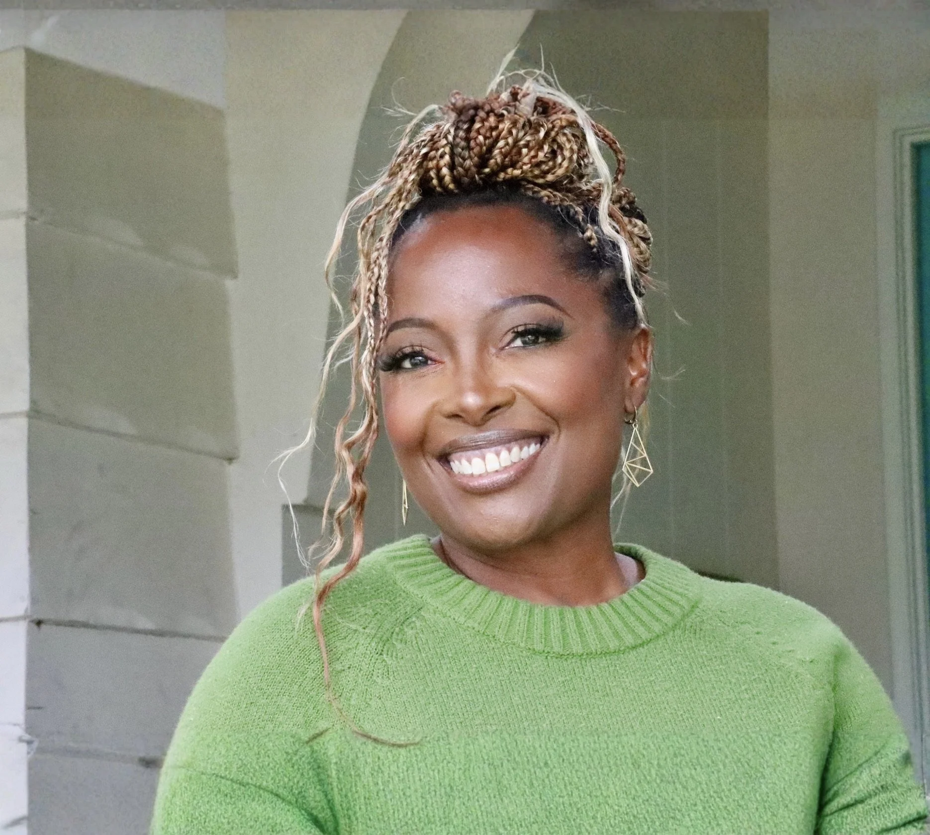 Close-up of a smiling woman with braided, blonde and brown hair styled in a high bun, wearing a bright green sweater and geometric gold earrings.