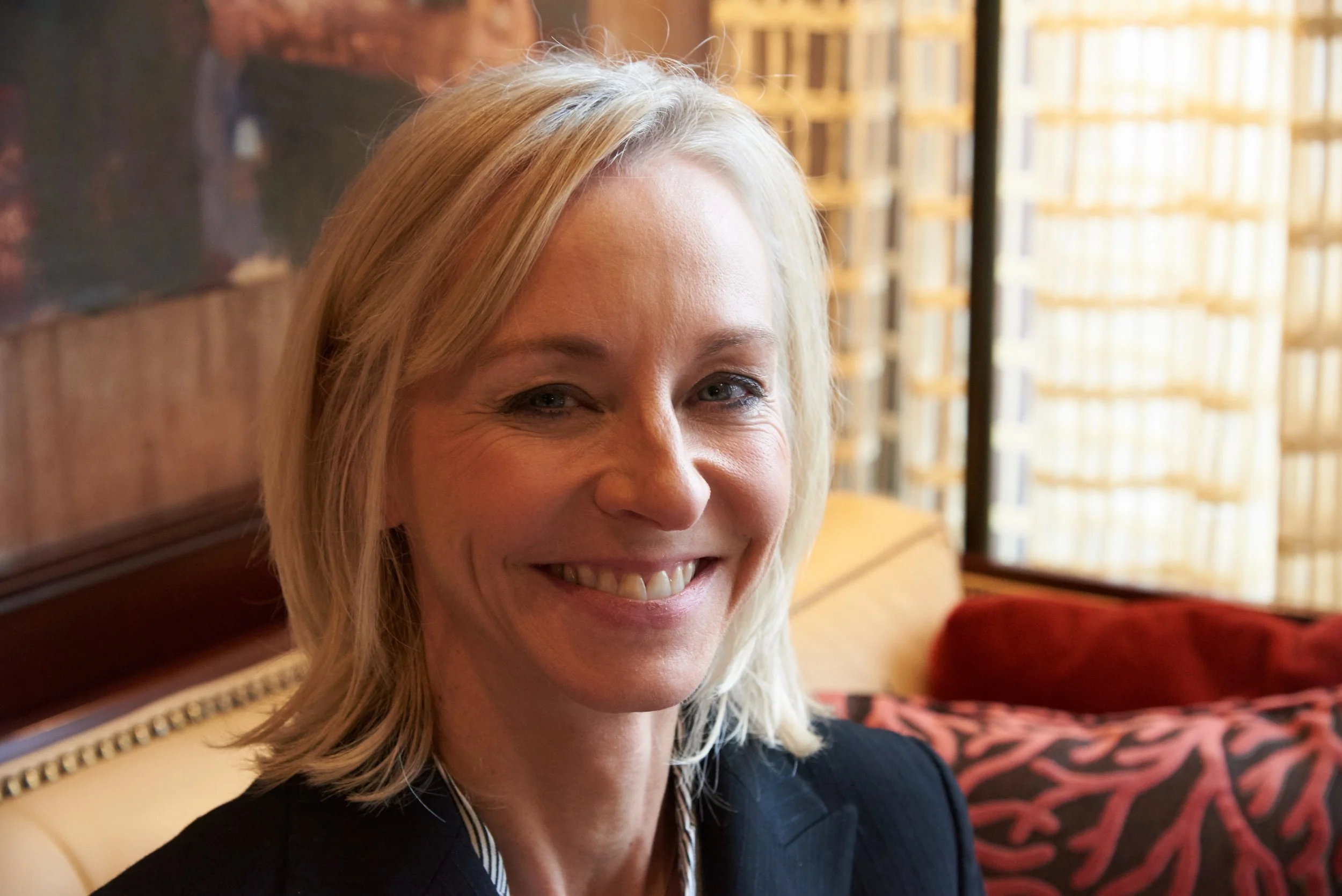 Close-up of a smiling blonde woman with blue eyes, wearing a black top, in an indoor setting with warm lighting, a wooden headboard, and patterned fabric in the background.