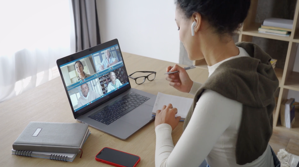 A woman in a white long-sleeve shirt and brown vest works at a desk during a video conference on her laptop, with four participants visible on the screen. She has wireless earbuds, a notebook, glasses, and a smartphone on the desk.
