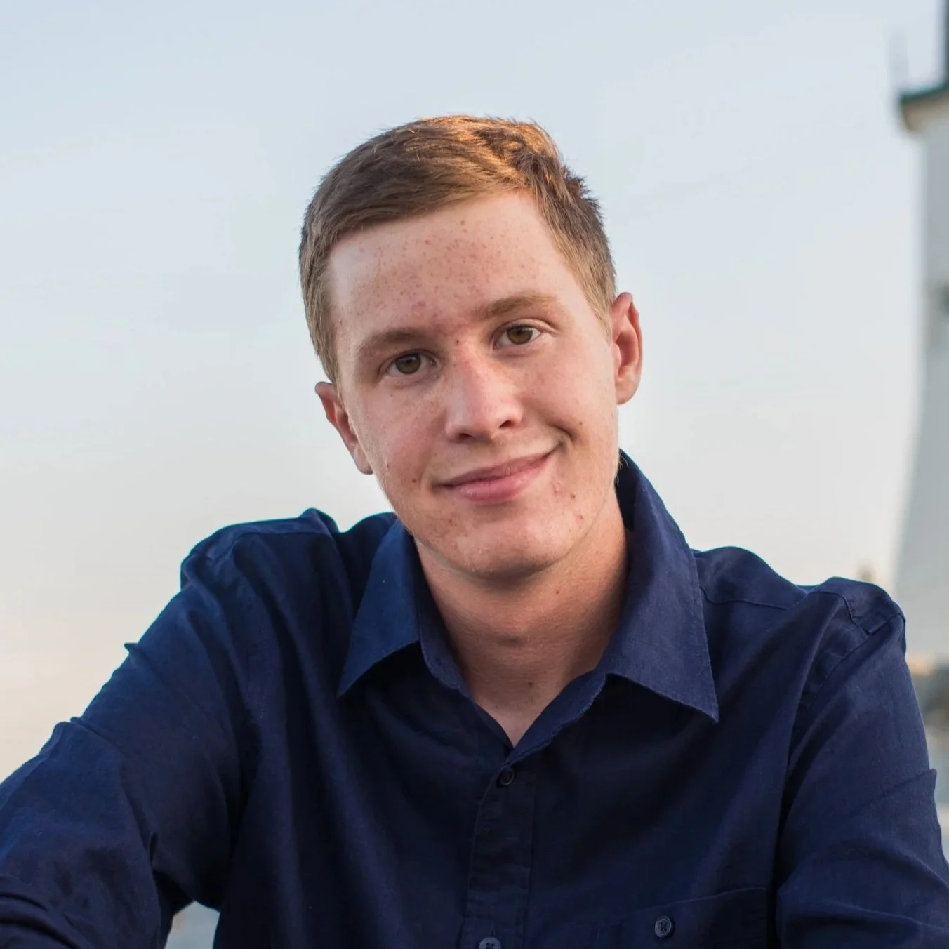 A young man with red hair and freckles wearing a navy blue shirt, smiling outdoors during the daytime.