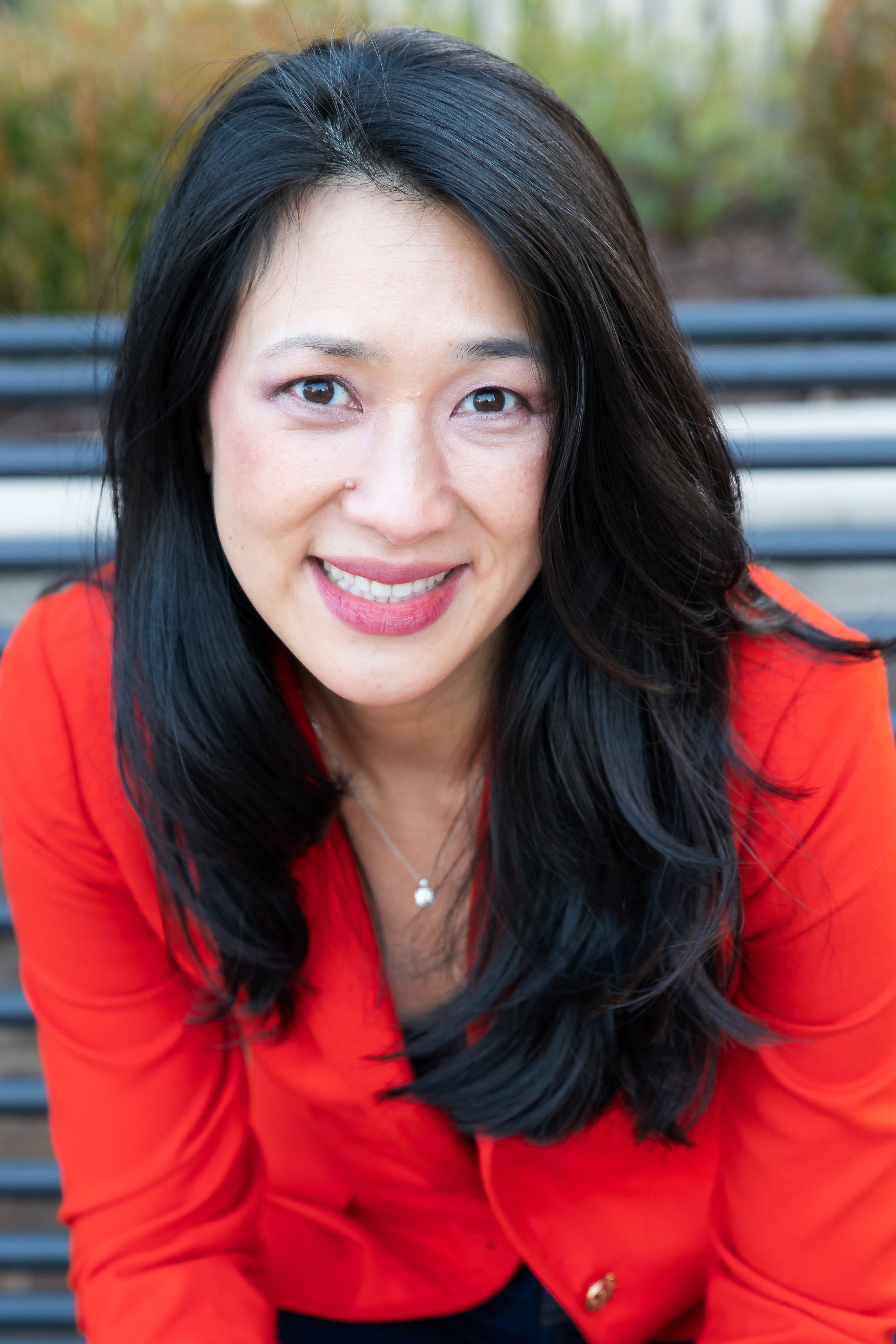 A woman with long black hair, wearing a red top, smiling at the camera outdoors with blurred green foliage in the background.