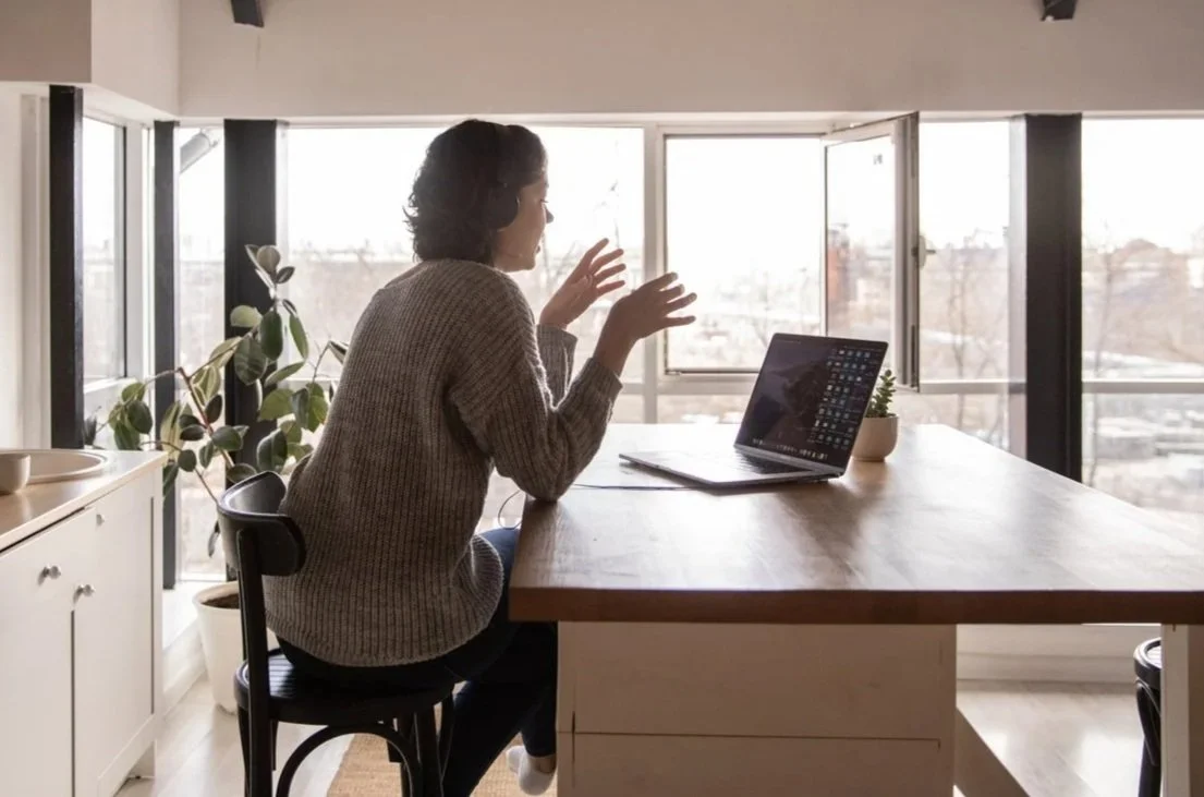 Woman sitting at a kitchen table talking on a video call, with a laptop, notebook, and potted plant, in a bright room with large open windows.