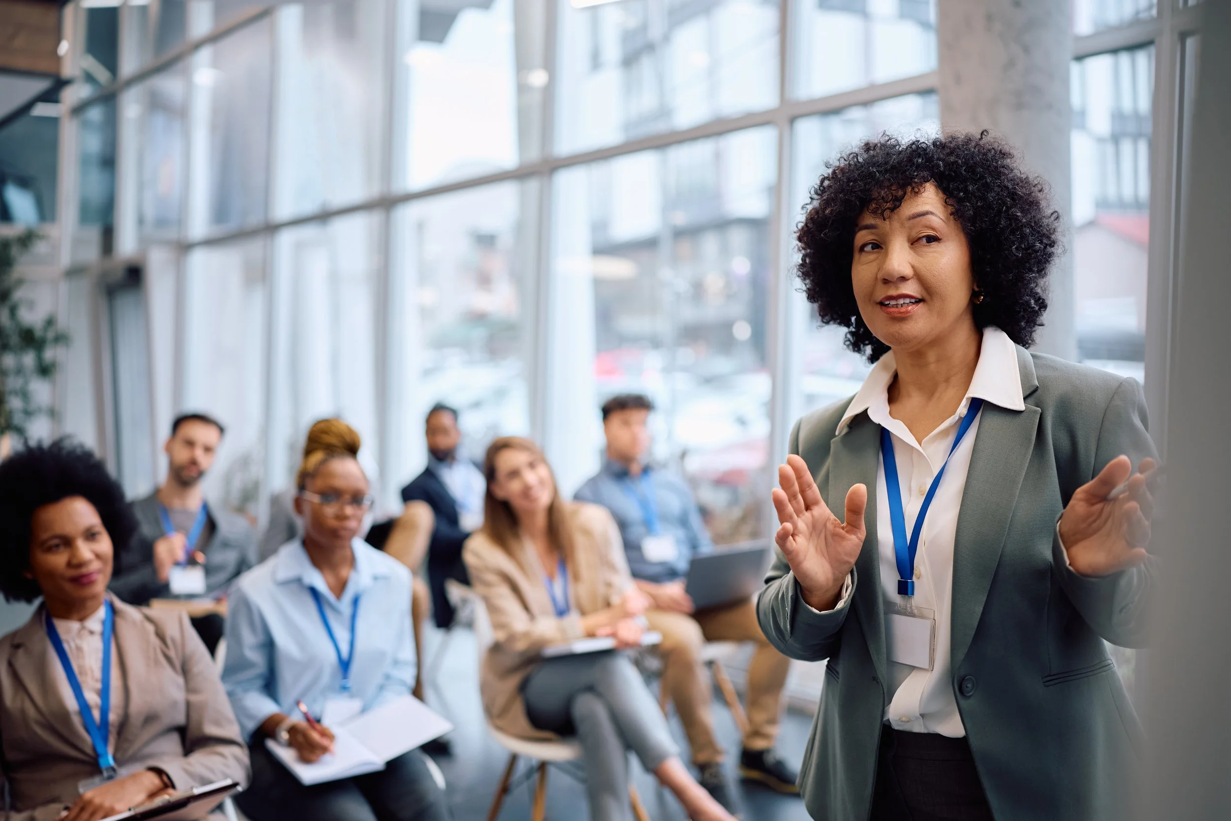 A woman in a gray blazer giving a presentation to a diverse group of people seated in a modern glass-walled conference room.