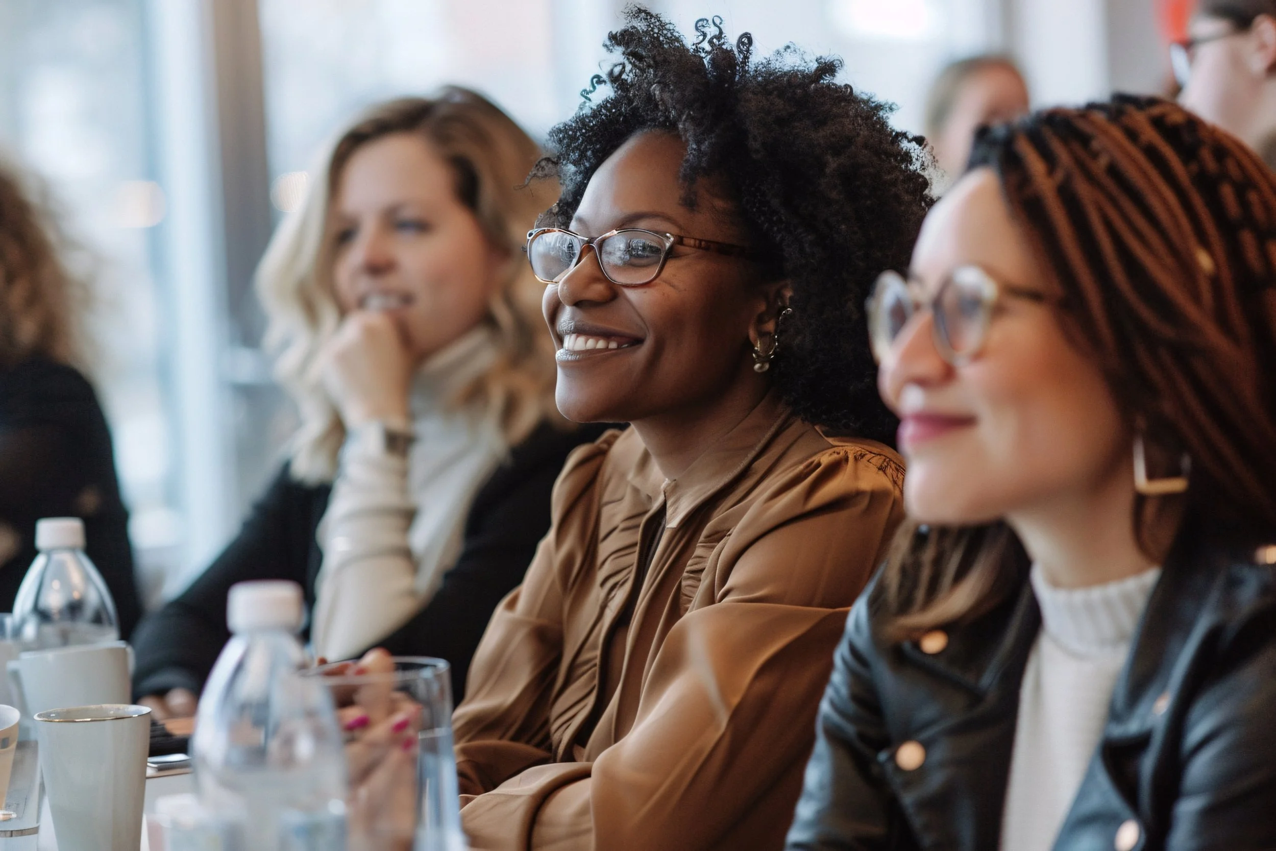 A group of women sitting at a conference table, smiling and listening attentively.