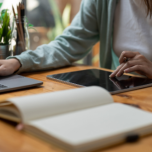 A person using a smartphone and working on a graphics tablet at a wooden desk with an open notebook and a closed laptop.