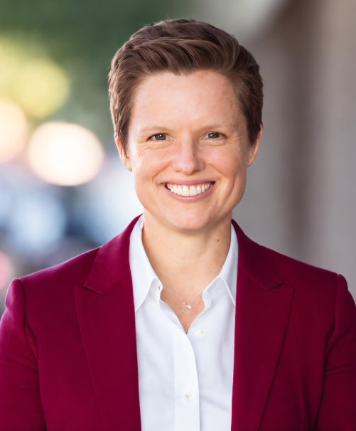 A woman with short brown hair wearing a white shirt and a maroon blazer, smiling outdoors with blurred trees and sunlight in the background.