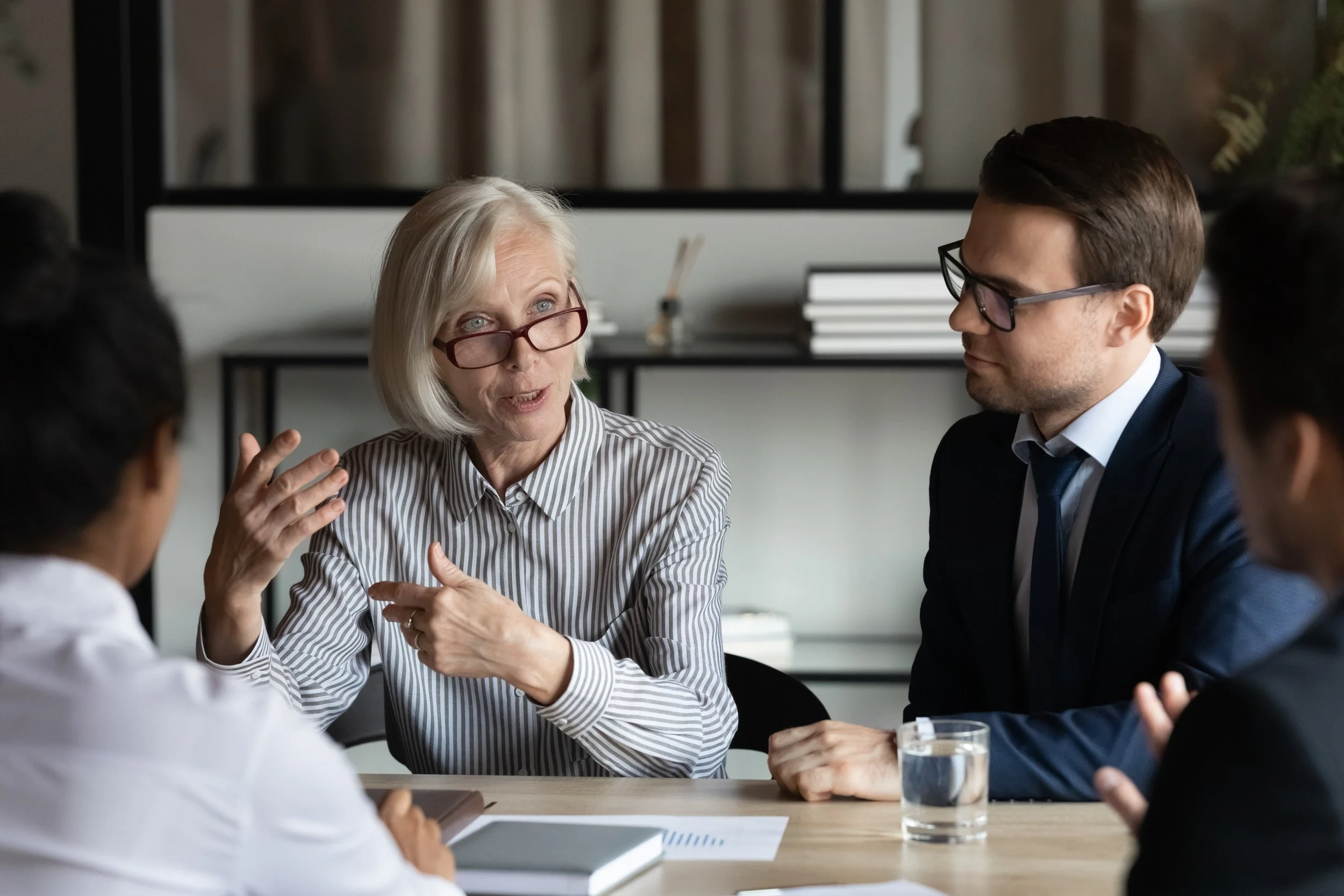 An elderly woman with glasses speaking passionately during a business meeting with three younger colleagues around a table.