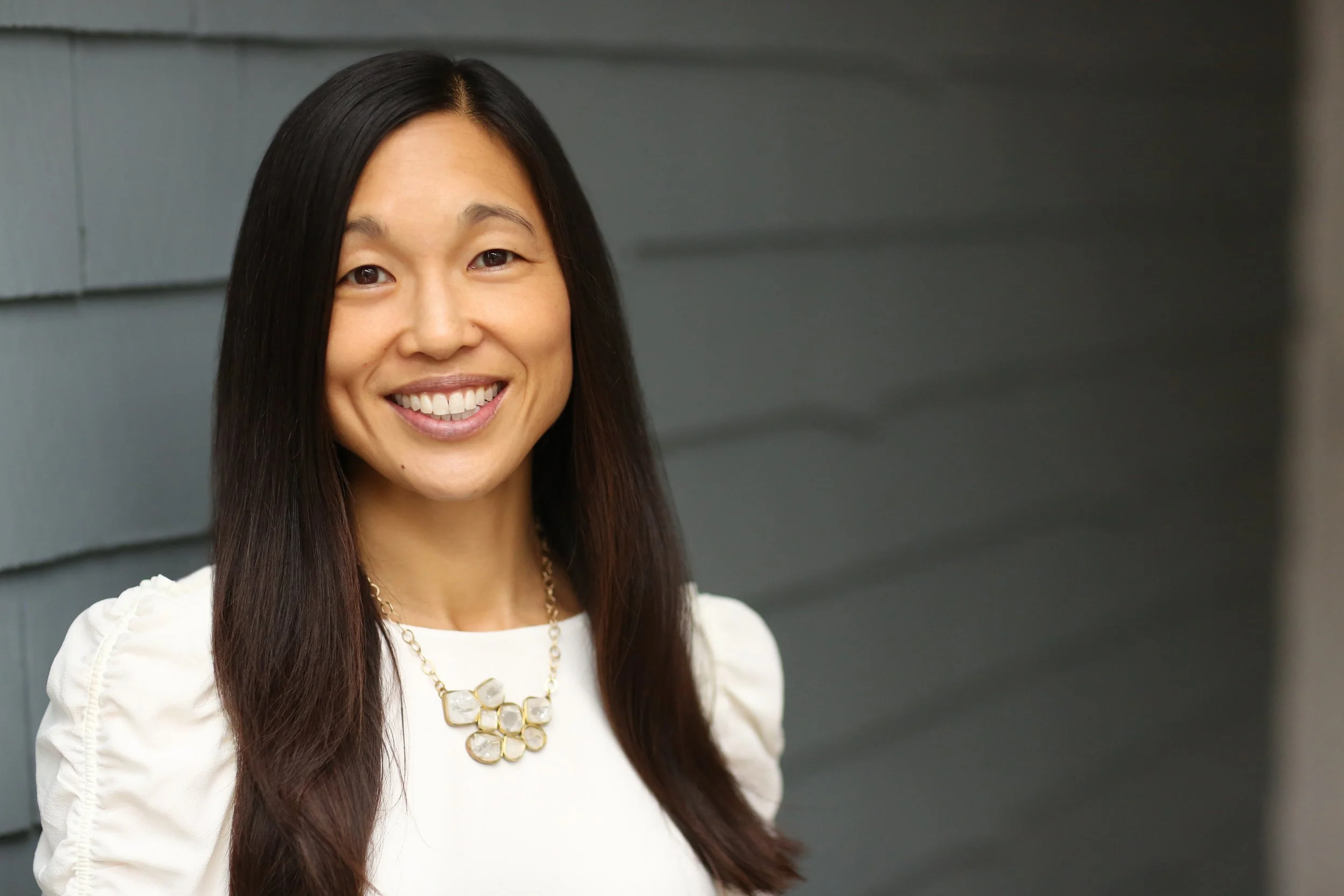 A woman smiling, wearing a white top and a statement necklace, with dark hair and standing against a gray wall.