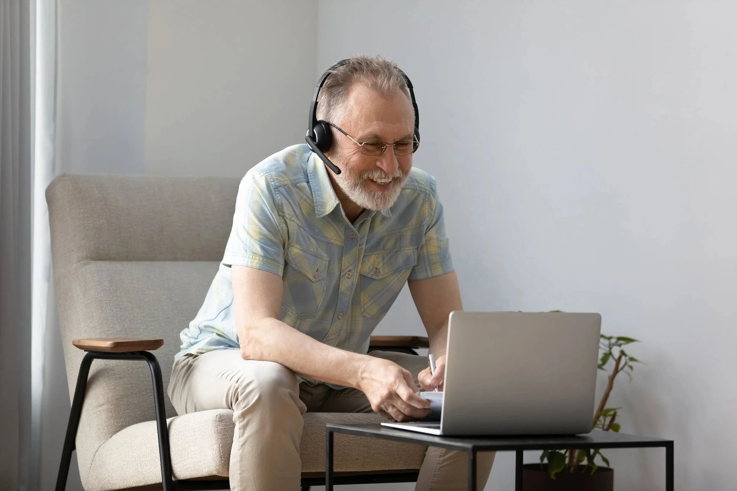 An older man with glasses and a beard, sitting on a beige sofa with a small black coffee table in front of him, wearing a headset, smiling while using a silver laptop.