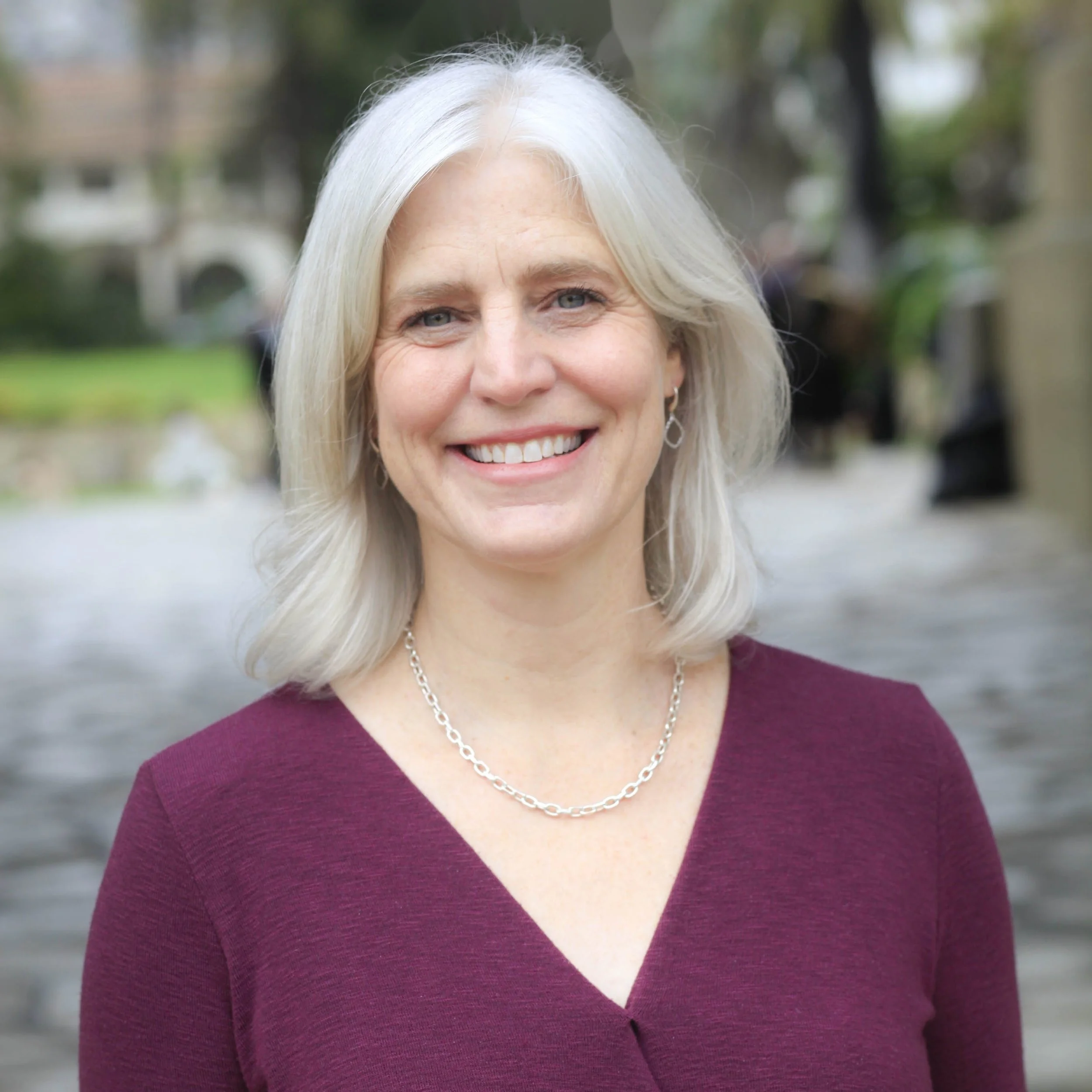 Close-up of a smiling woman with long white hair, wearing a purple top and silver necklace, outdoors with blurred trees and buildings in the background.