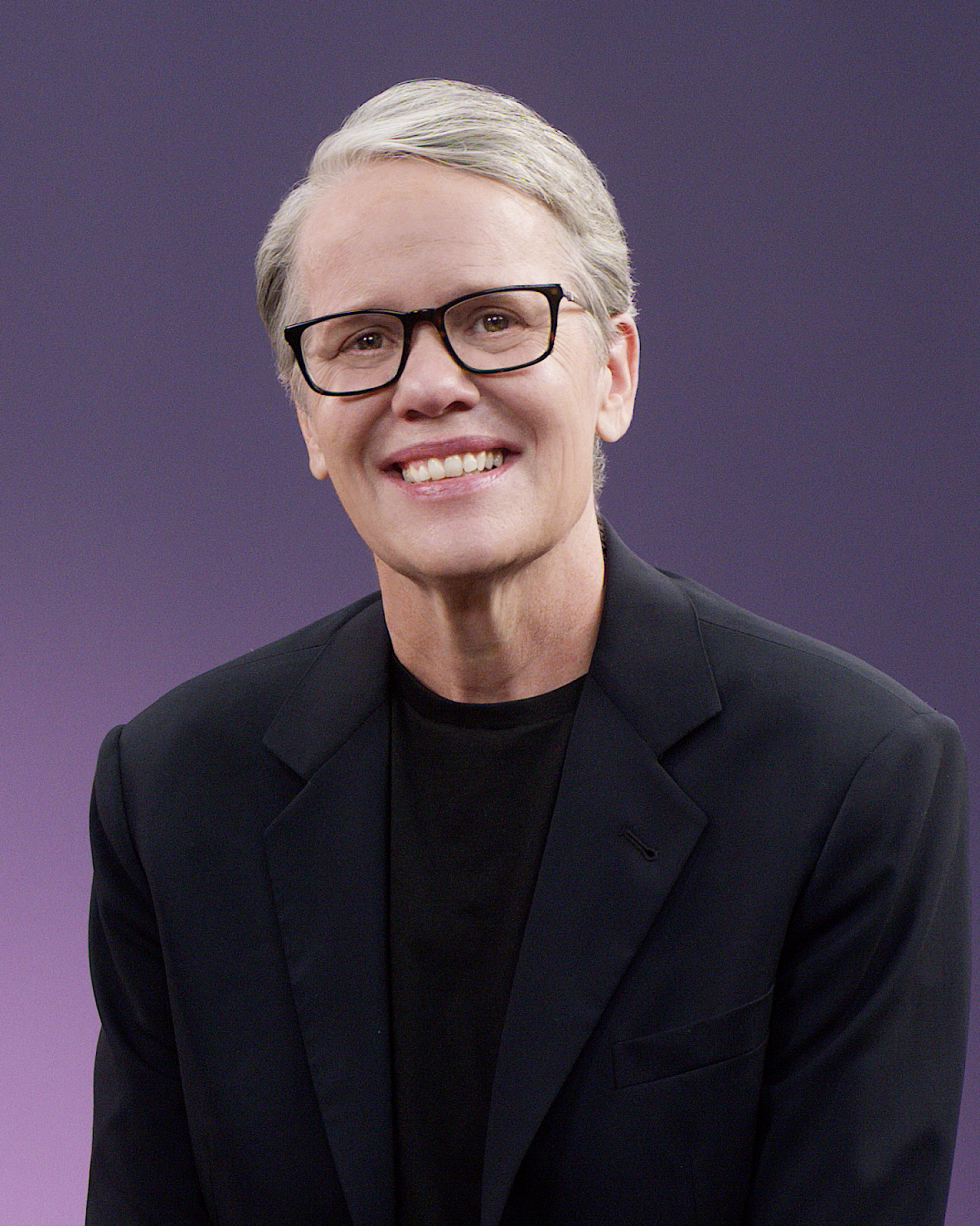 Portrait of a woman with short gray hair, black glasses, wearing a black blazer and black shirt, smiling against a purple background.