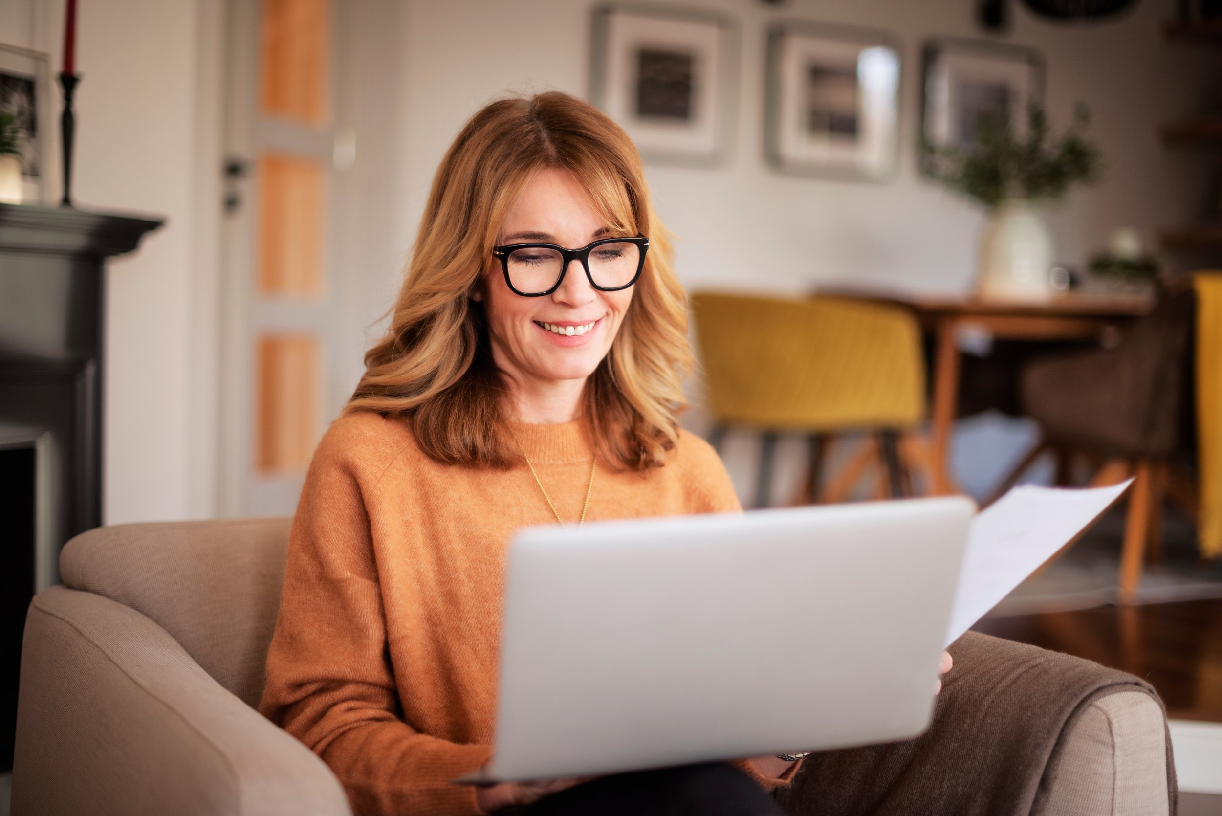 A woman with light brown hair, glasses, and an orange sweater sitting on a beige couch, smiling while looking at a laptop with papers in her hand, in a cozy living room with framed pictures and a table with chairs in the background.