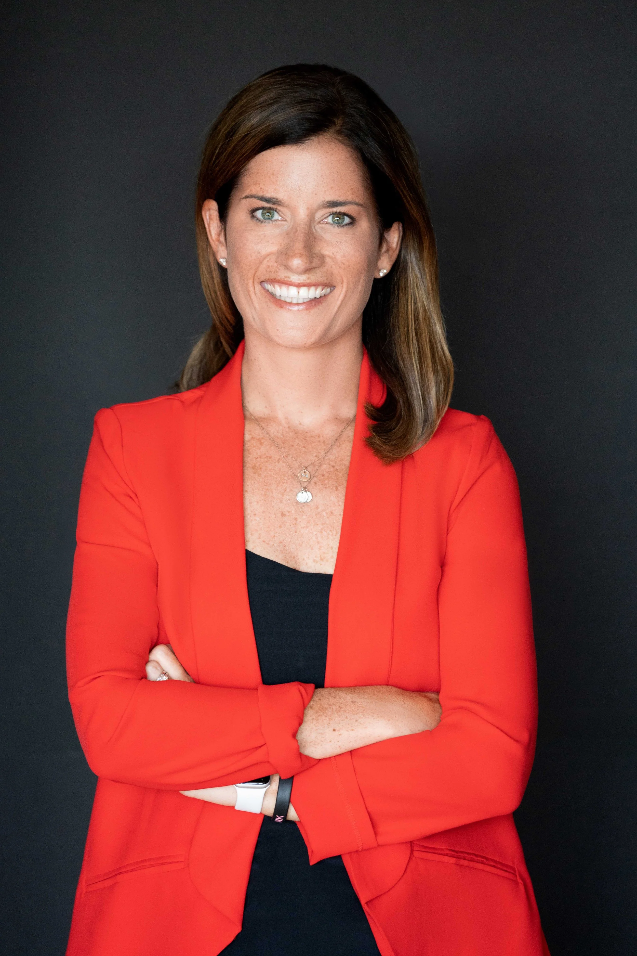 A woman with brown hair, green eyes, and freckles, smiling with arms crossed, wearing a red blazer over a black top, against a dark background.