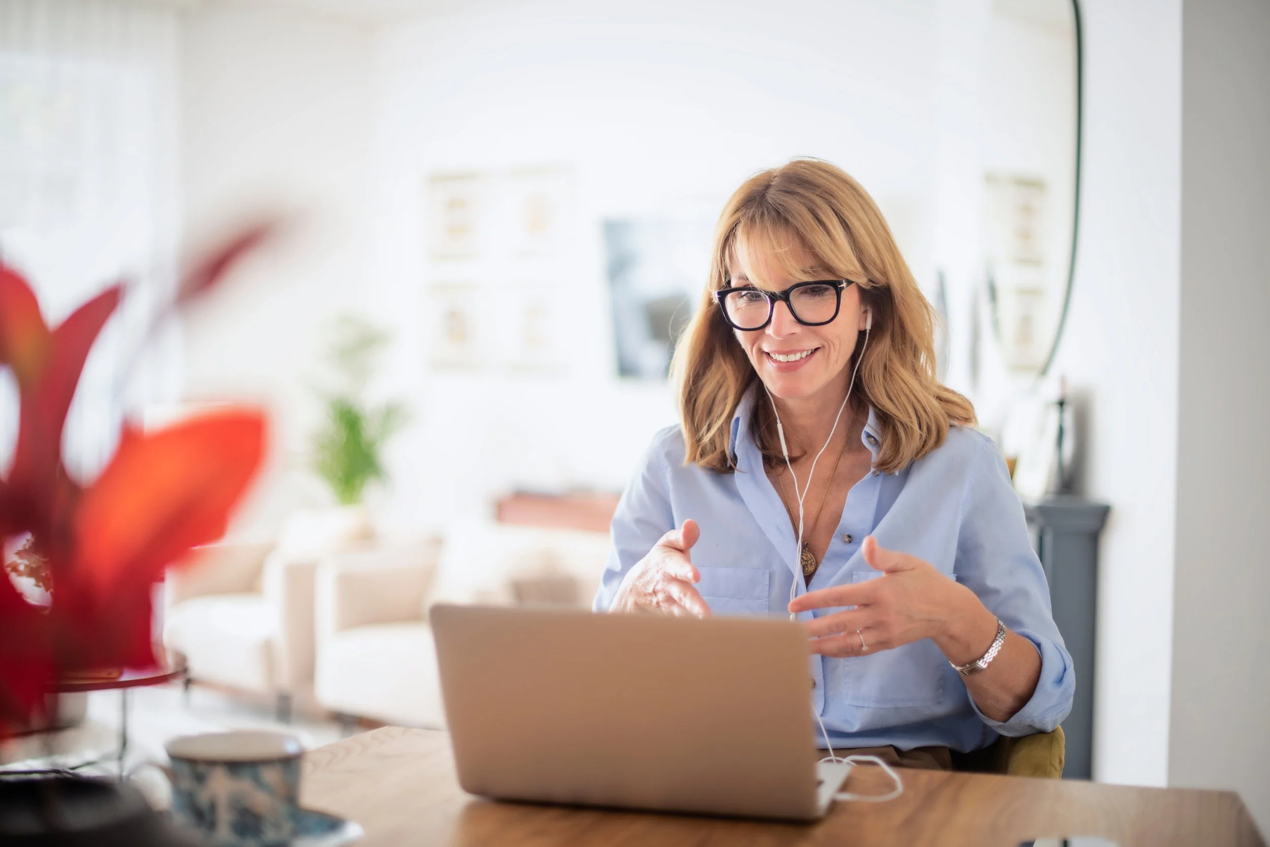 A woman with glasses and shoulder-length hair smiling while on a video call, sitting at a wooden table with a laptop, wearing earphones, in a bright room