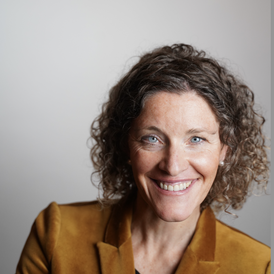 A woman with curly brown hair and blue eyes smiling, wearing a mustard-colored jacket, against a plain neutral background.