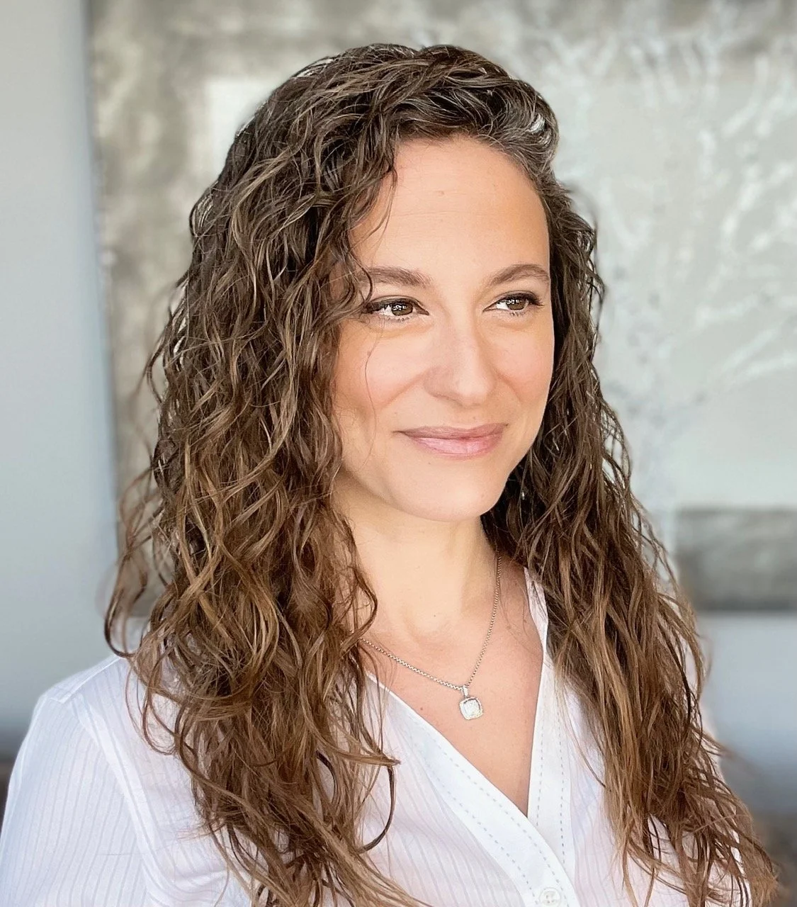 Portrait of a smiling woman with curly hair wearing a teal top with a decorative necklace, standing against a wooden background.