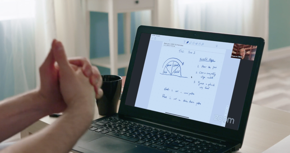 A person on a video call with a presentation on a laptop screen. The presentation includes a pie chart and handwritten notes. The person's hands are clasped together.