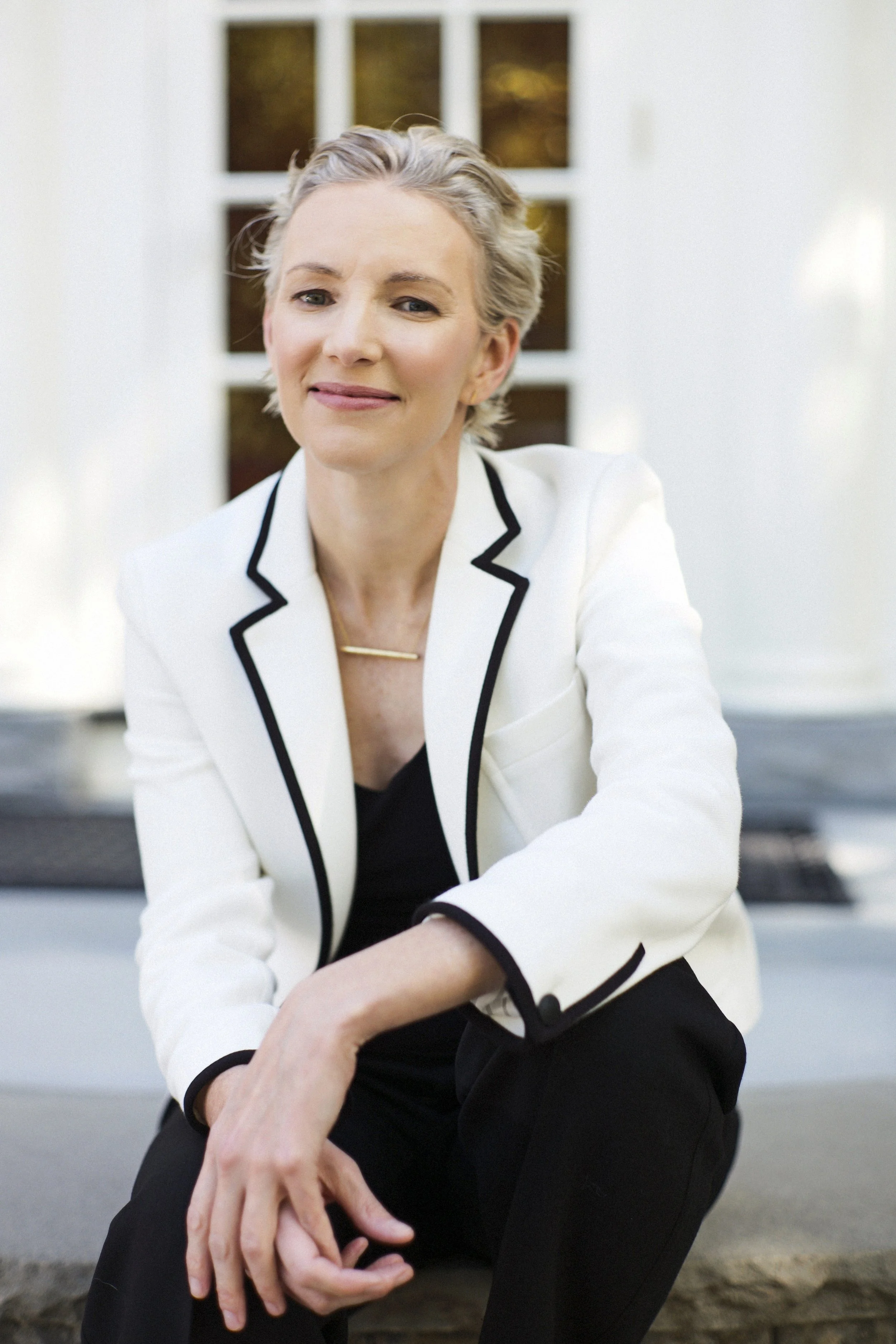 A woman with short gray hair wearing a white blazer with black trim, sitting outdoors in front of a house with white siding and windows, smiling at the camera.