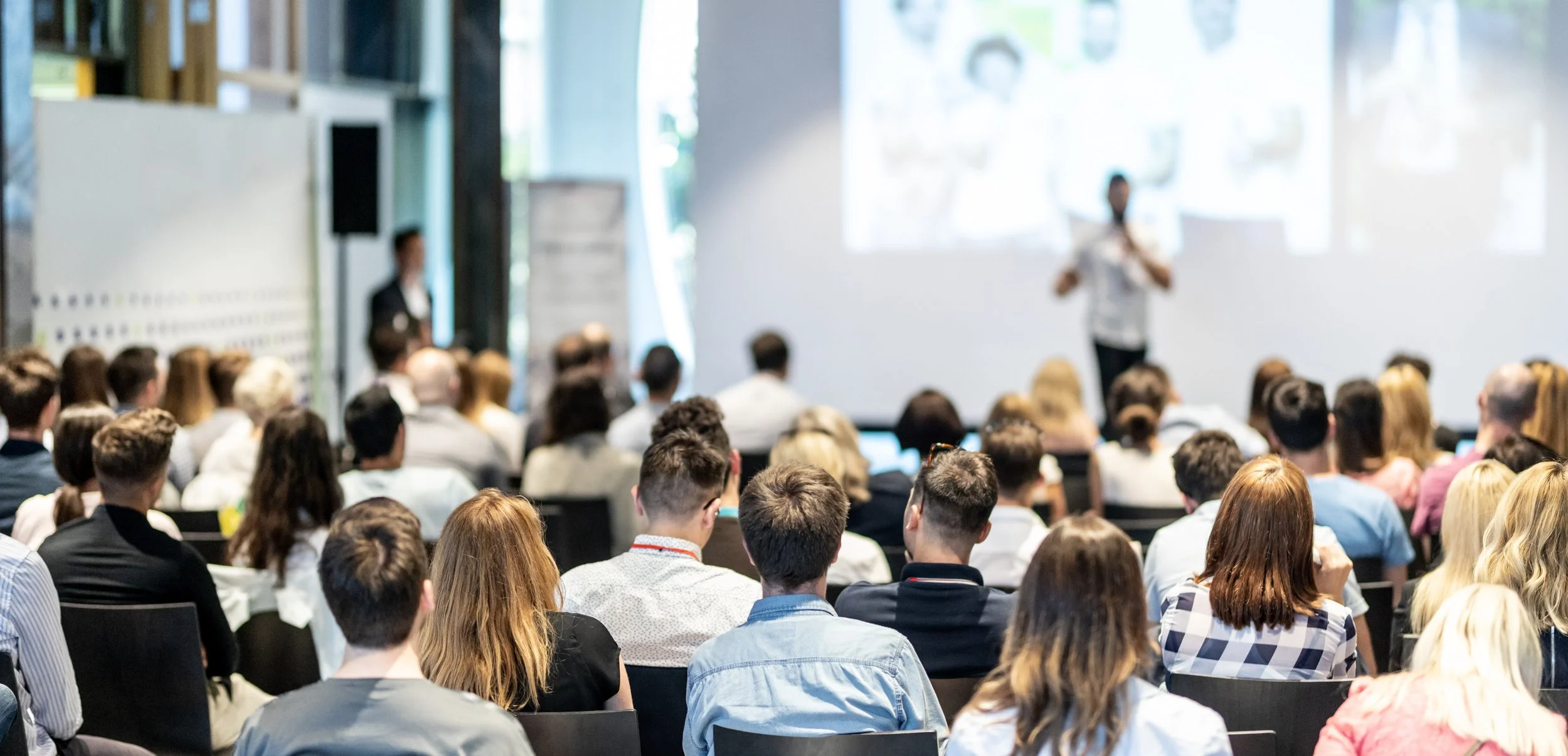 A large group of people attending a conference or seminar, facing a presenter at the front of the room with a large screen behind them.