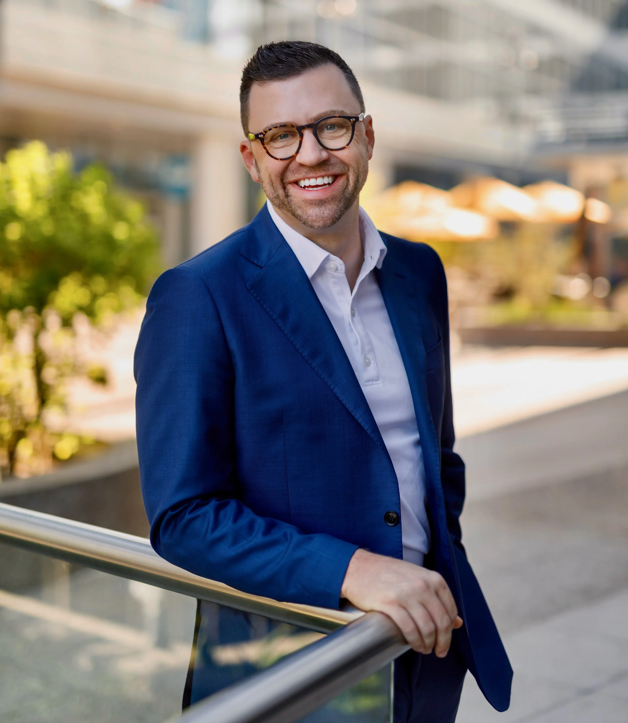 A man in a blue suit and glasses smiling outdoors on a sunny day.