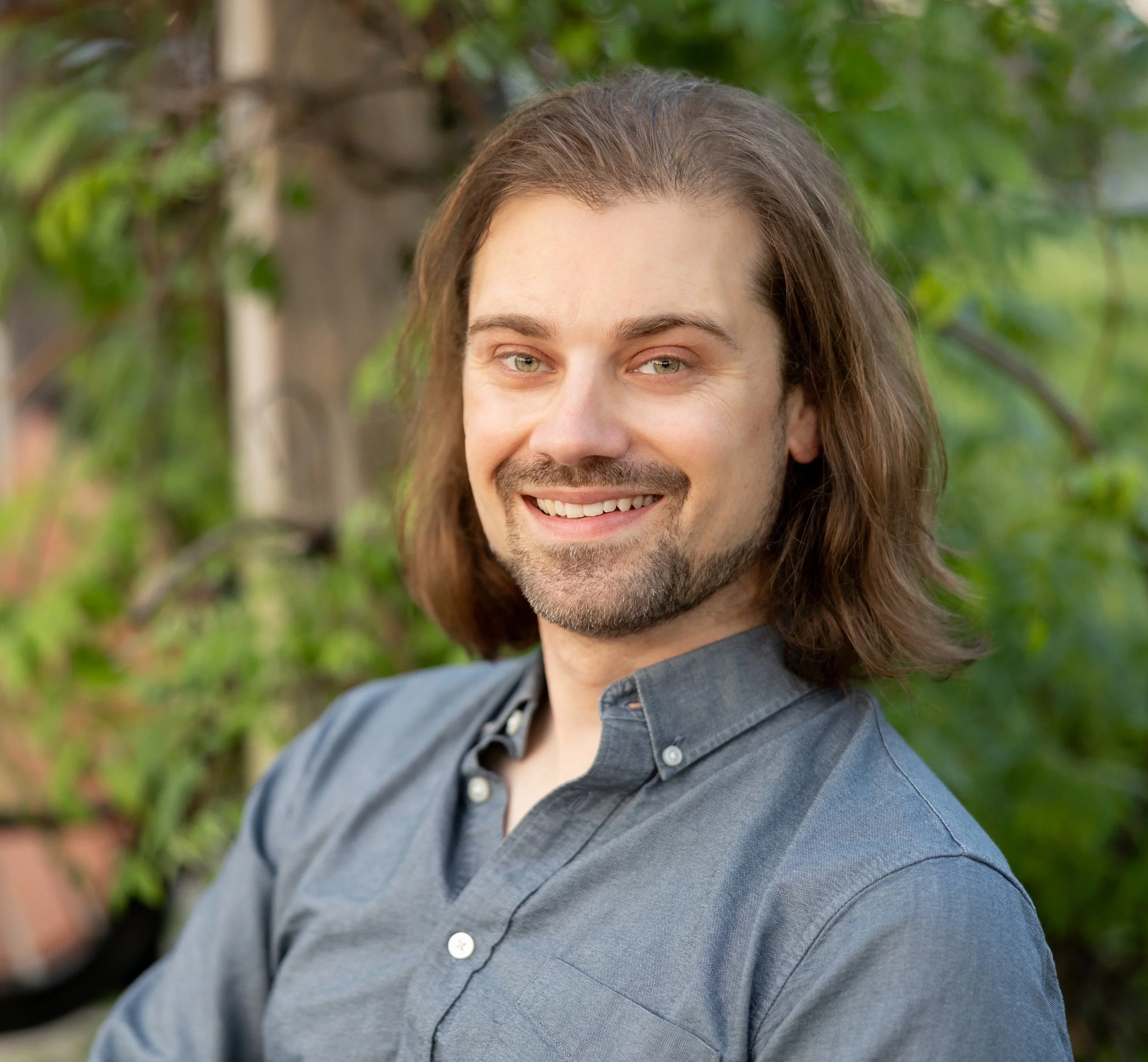 Close-up of a man with shoulder-length brown hair and a short beard, smiling and wearing a gray button-up shirt, outdoors with blurred green foliage in the background.