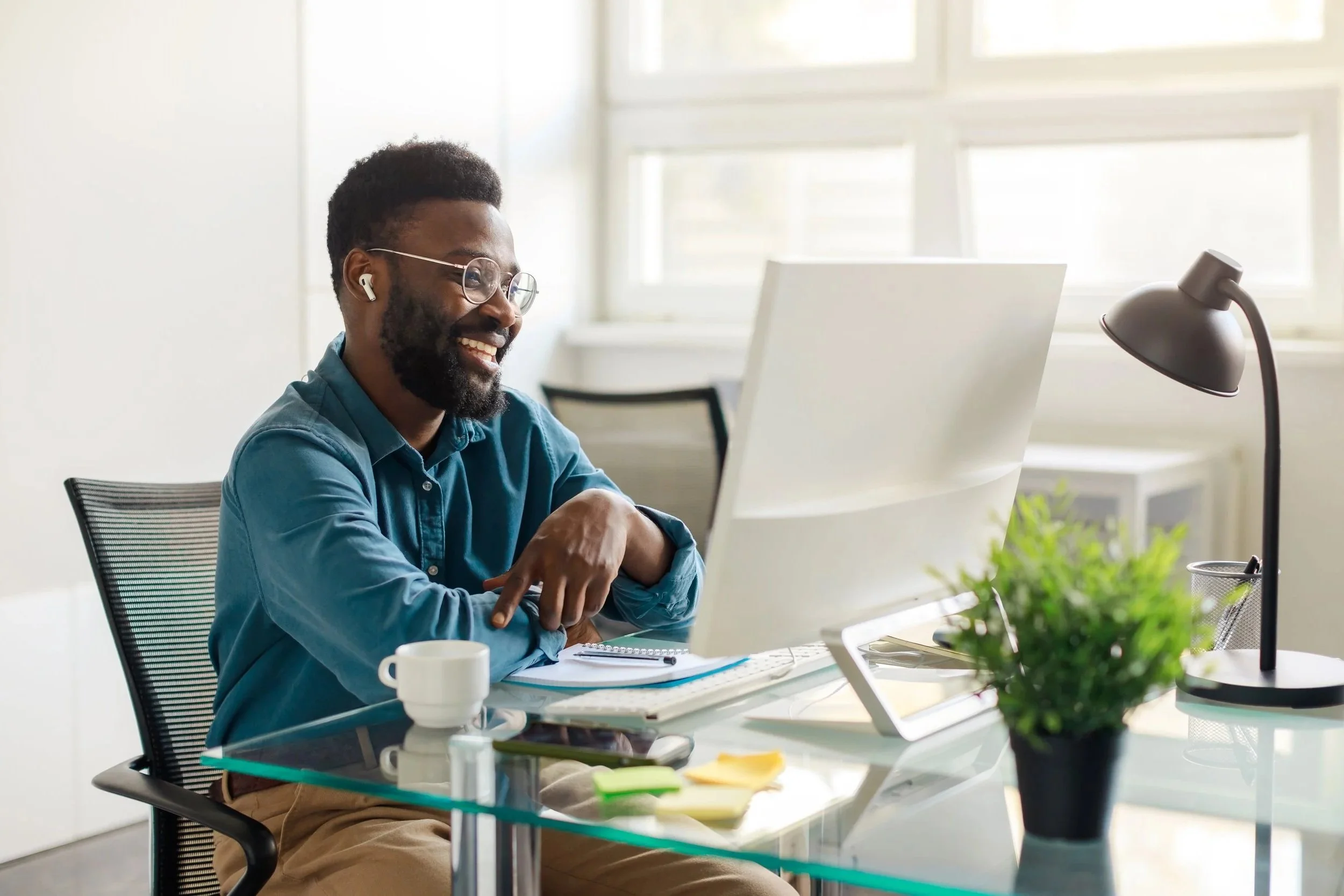 Man smiling at a computer screen in an office, wearing glasses and wireless earbuds, surrounded by a coffee cup, notebook, and plant.