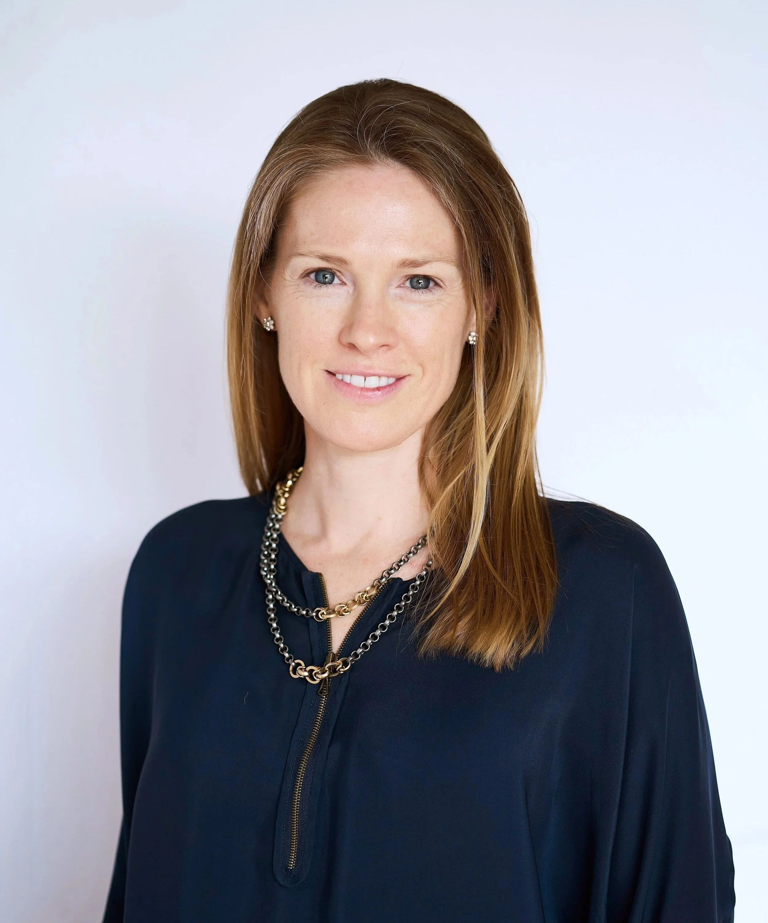 A woman with red hair wearing a black top and gold and silver jewelry, smiling at the camera against a white background.