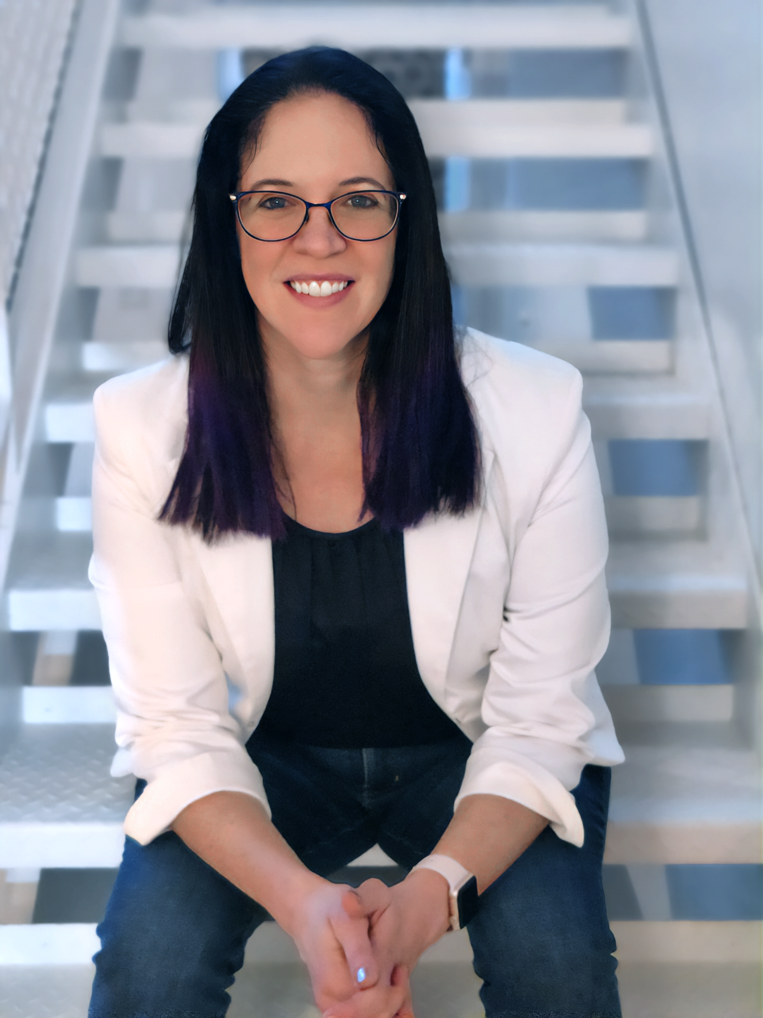 A woman with black hair and purple-dyed tips, wearing glasses, a white blazer, and a black top, sitting on a staircase, smiling at the camera.