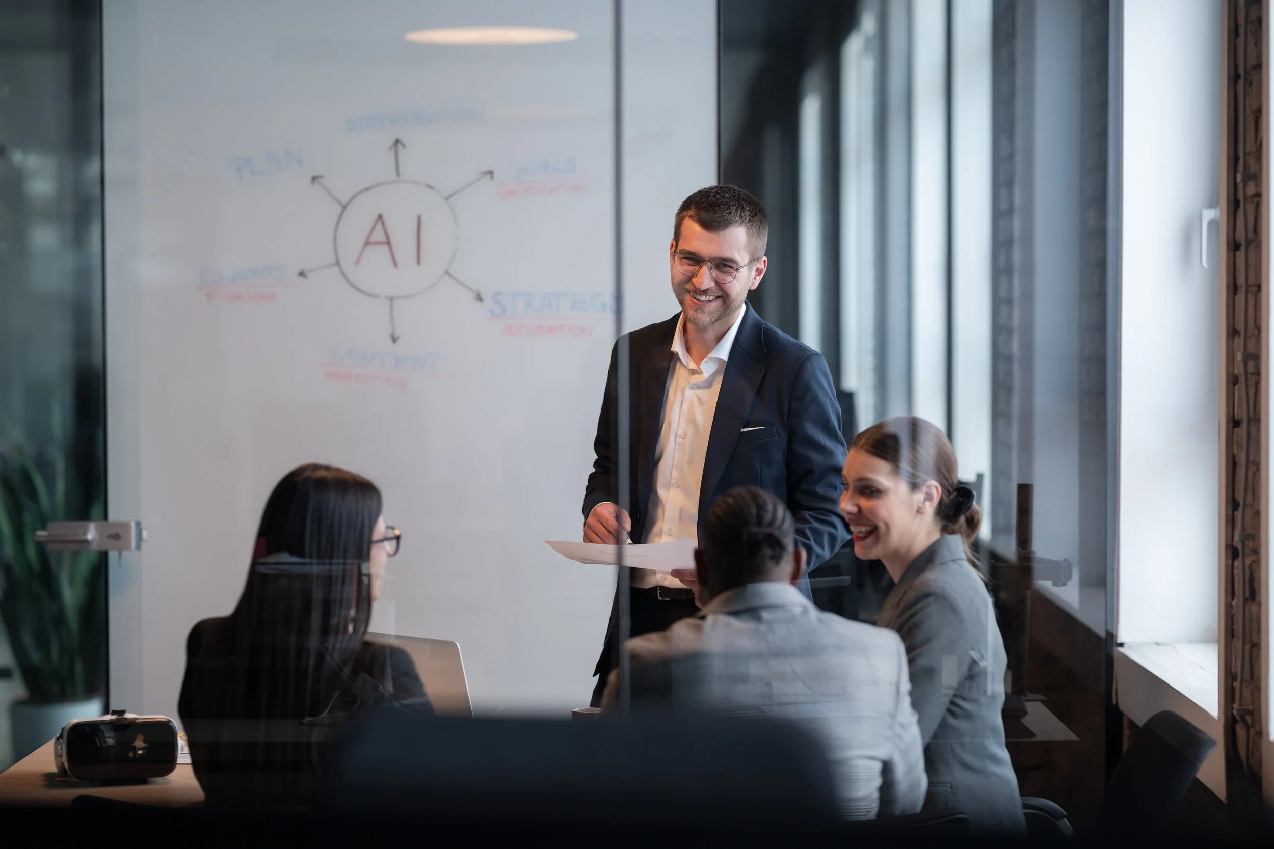 A man in a suit presenting to four women in a meeting room, with a whiteboard showing a diagram of 'AI' and related terms in the background.