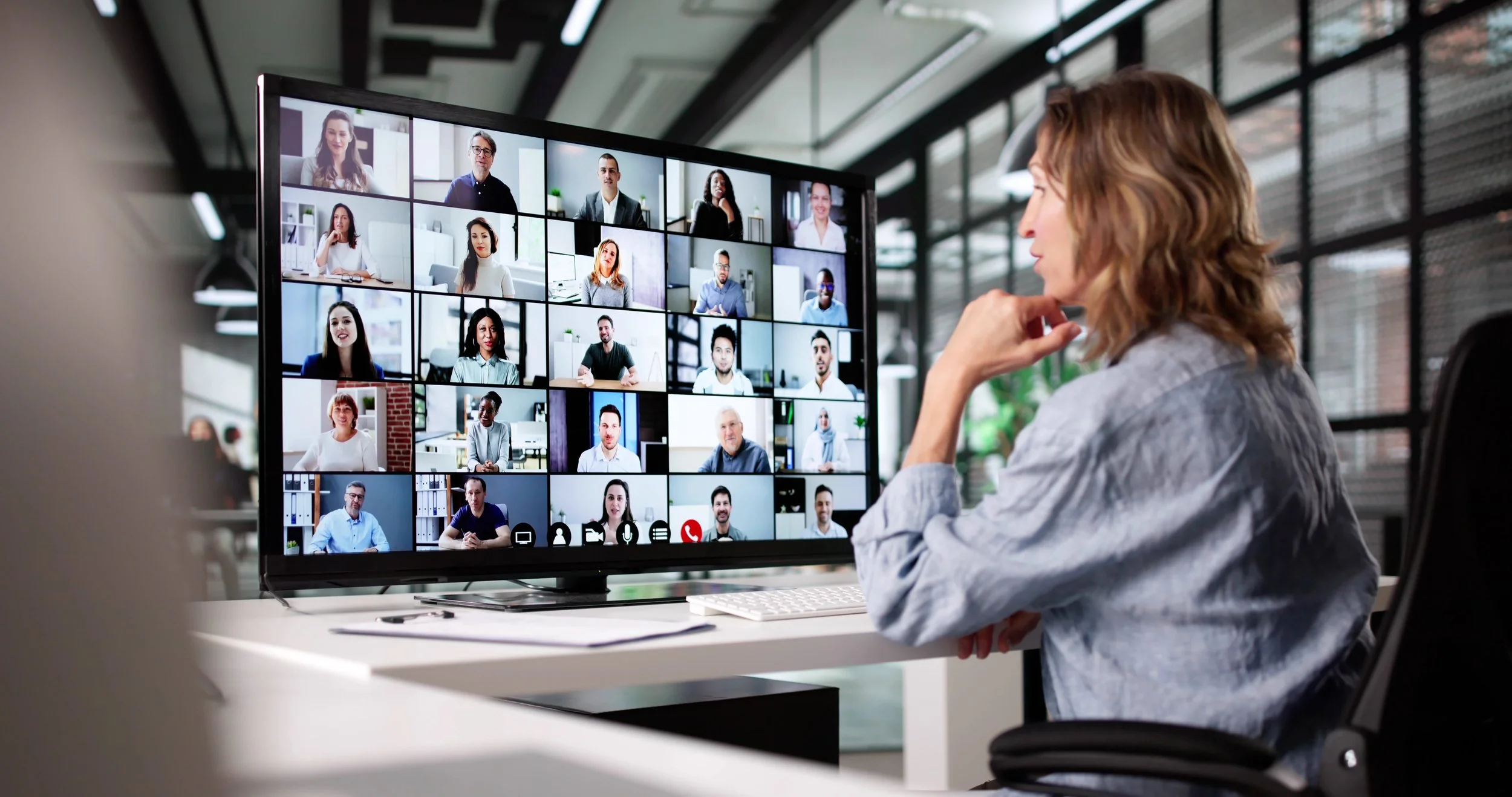 Woman participating in a virtual video conference call with multiple people displayed on a large screen in an office setting.
