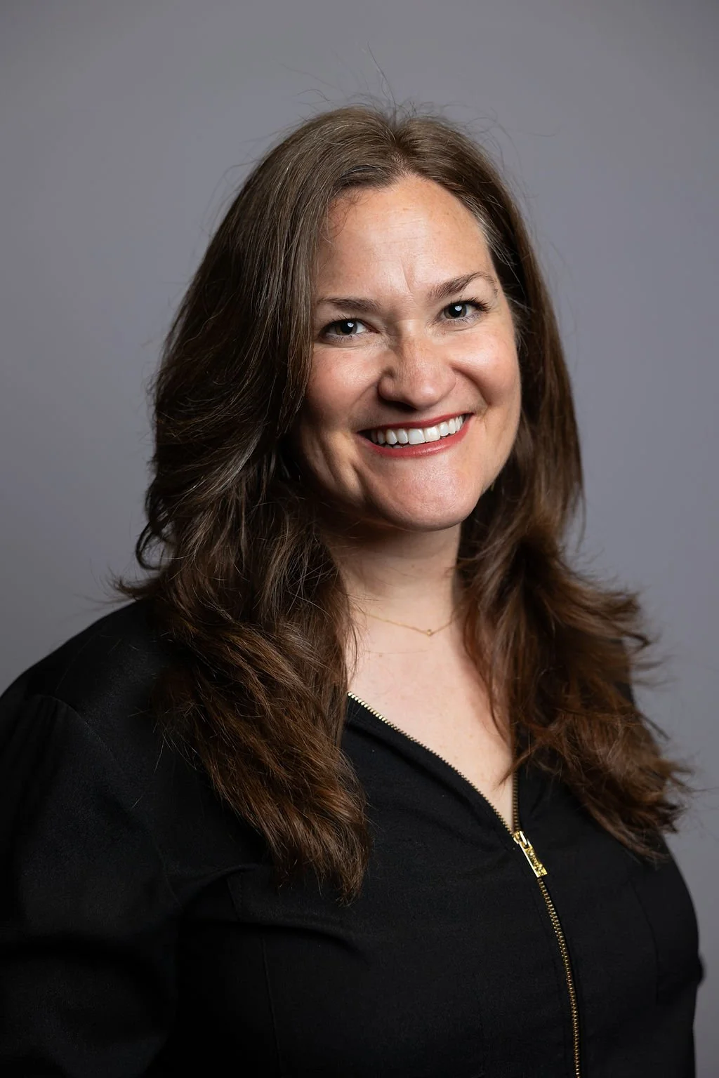 A woman with long brown hair, smiling, wearing a black top with a zipper, and a small gold necklace, standing against a gray background.