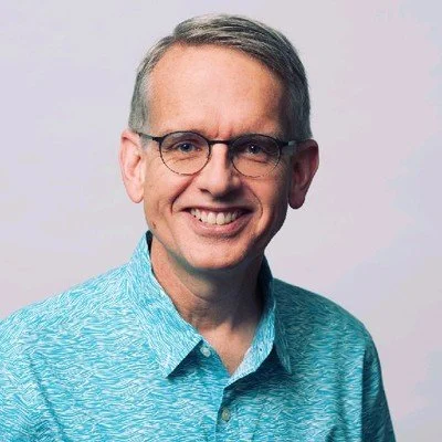 A smiling middle-aged man with glasses wearing a blue patterned collared shirt, posing against a plain light background.