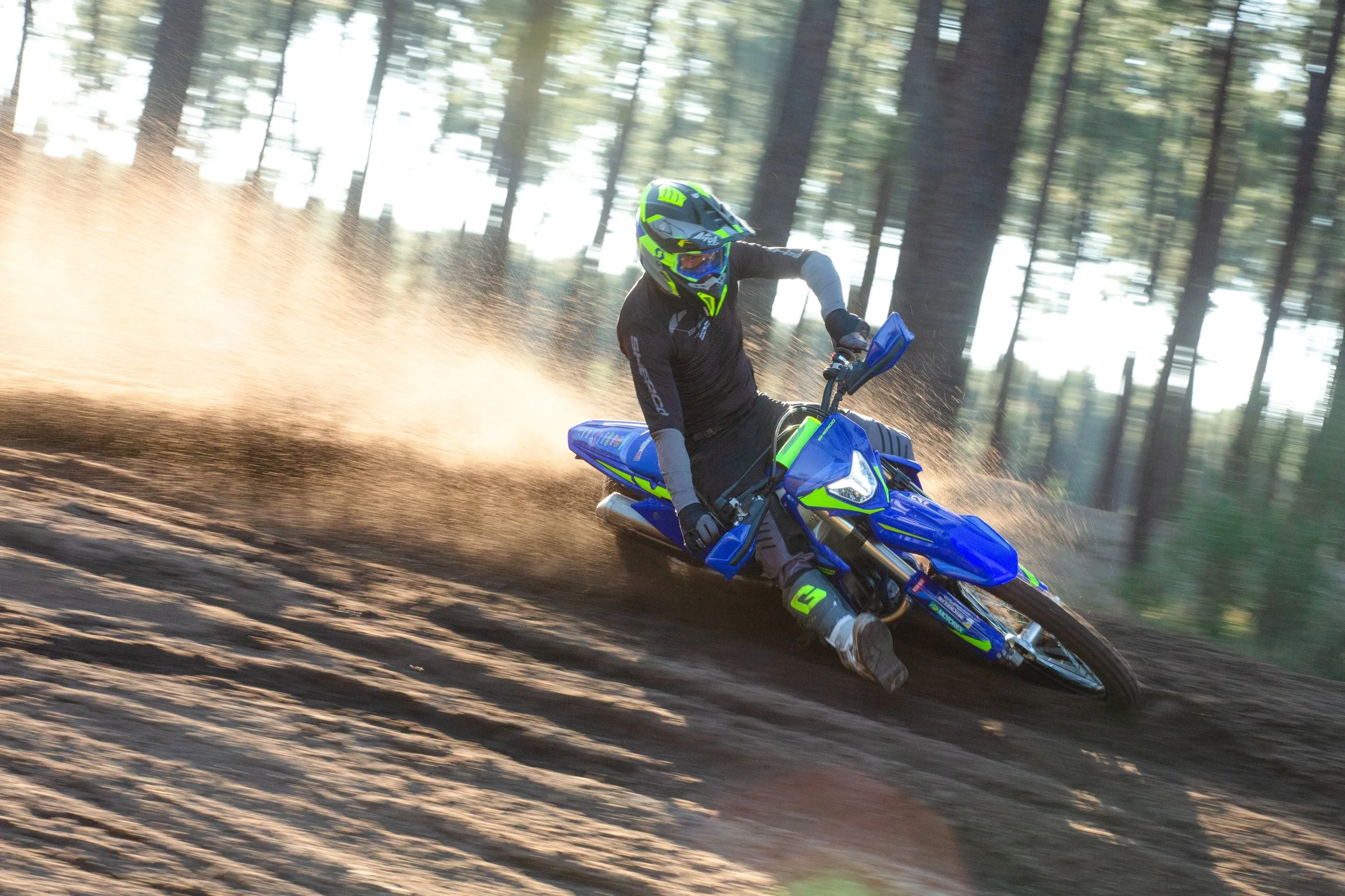 Sherco dirt bike rider in black and neon green gear navigating a turn on a dirt trail, kicking up dust in a forested area.