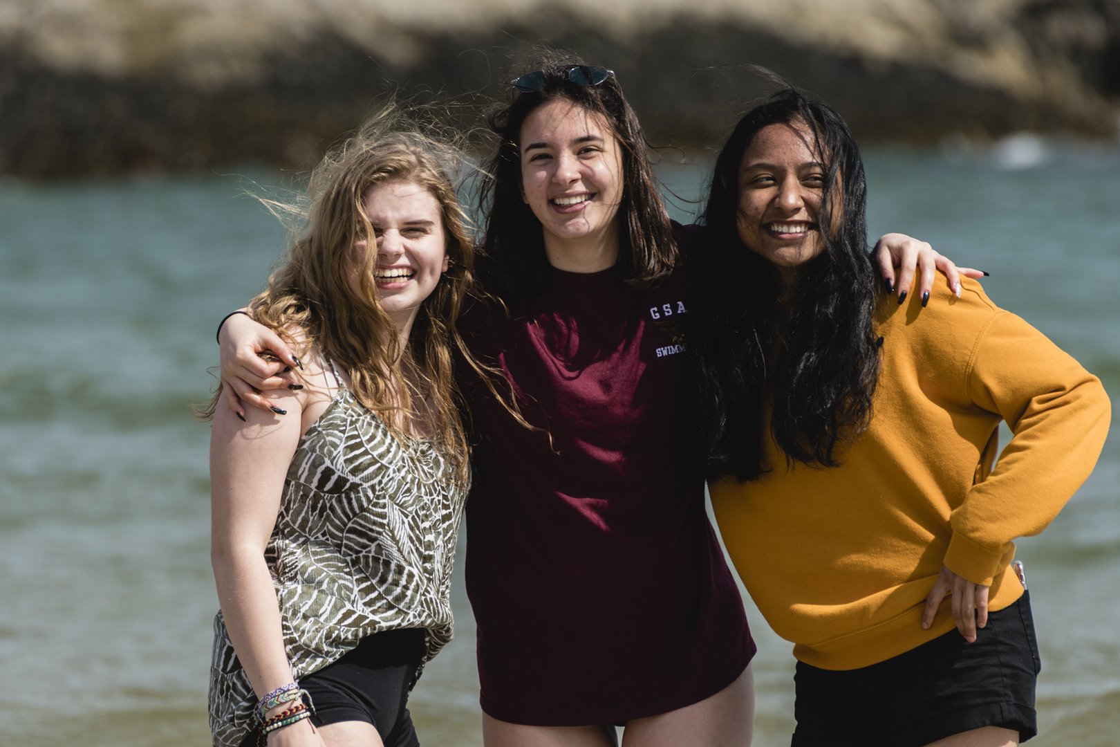 Three smiling young women embracing each other on a beach with clear water in the background.