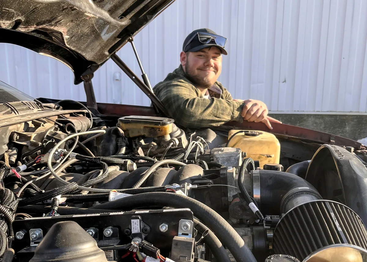 A man is sitting inside an open car hood, smiling and leaning on the car's engine. The engine is exposed, showing various components, wires, and parts. The background features a white wall, and the man is wearing a plaid shirt, sunglasses, and a cap.