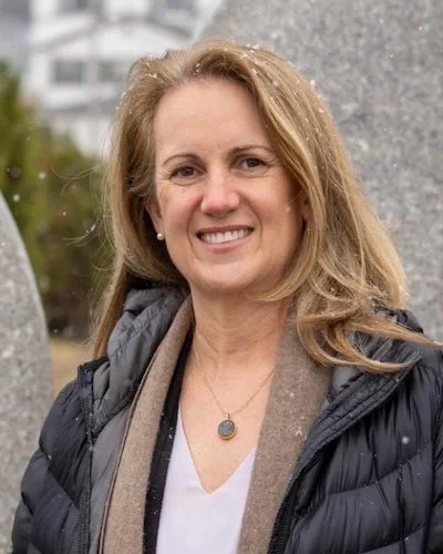 Portrait of a woman with shoulder-length blonde hair, smiling, wearing a dark top and earrings, against a gray background.