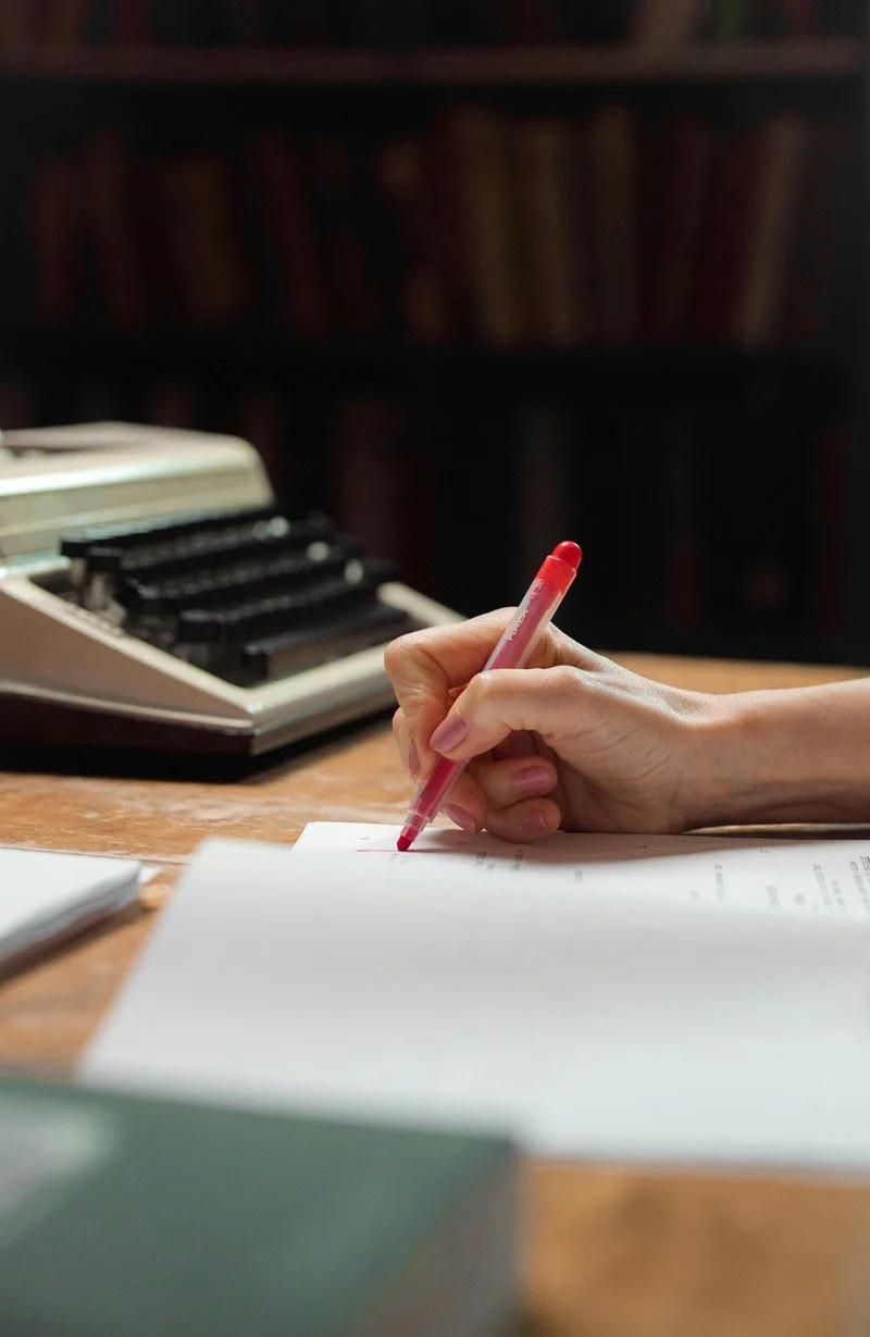 Alt text: Hand holding red pen over printed pages, with a typewriter in the background.
