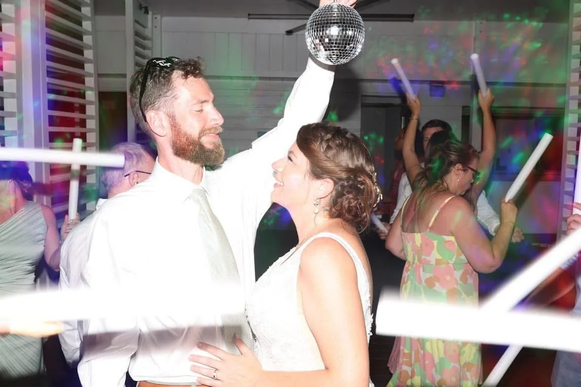 A wedding reception with guests dancing, including a man with a beard holding a disco ball and dancing with a woman in a white dress.
