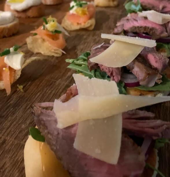 Close-up of a wooden serving board with assorted small beef and seafood appetizers topped with cheeses and garnishes.