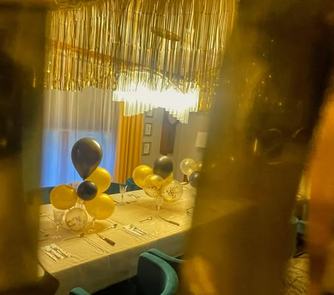 Decorated dining table with yellow, black, and clear balloons in a dimly lit room with a chandelier and curtains.